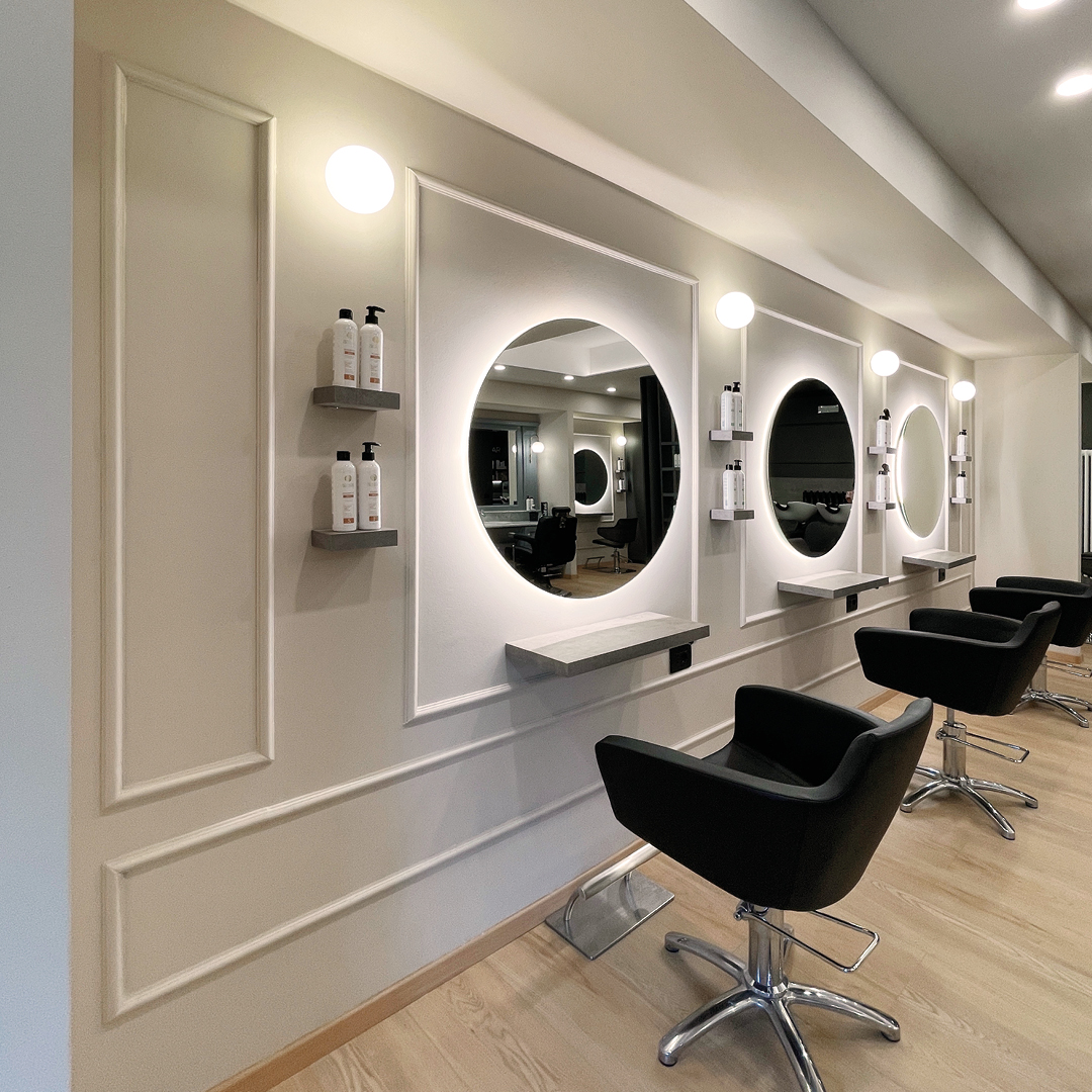 Focus su una postazione dell'area stilistica con pareti colo panna e dettagli decorativi in rilievo in evidenza. Poltrona nera e specchio rotondo contornato da luci led fredde decorative 